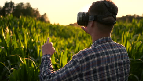 A-modern-farmer-in-a-VR-helmet-drives-a-corn-crop-standing-in-a-field-at-sunset-in-the-sun.-The-concept-of-smart-fields-of-use-of-neural-networks-in-agriculture.-The-use-of-artificial-intelligence-to-agriculture-and-harvest